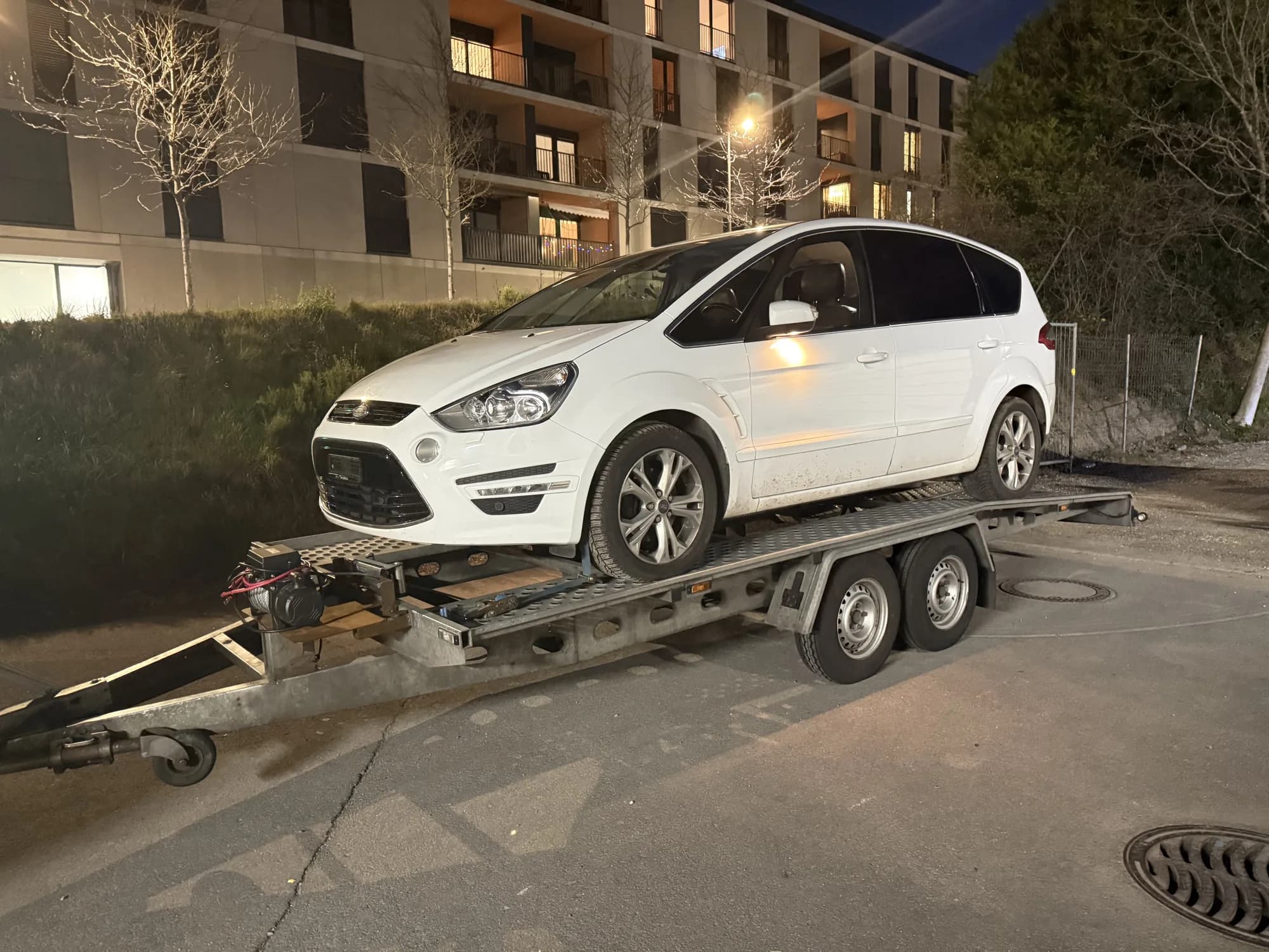 Monospace blanc chargé sur une remorque porte-voiture à double essieu, de nuit, devant un immeuble résidentiel éclairé