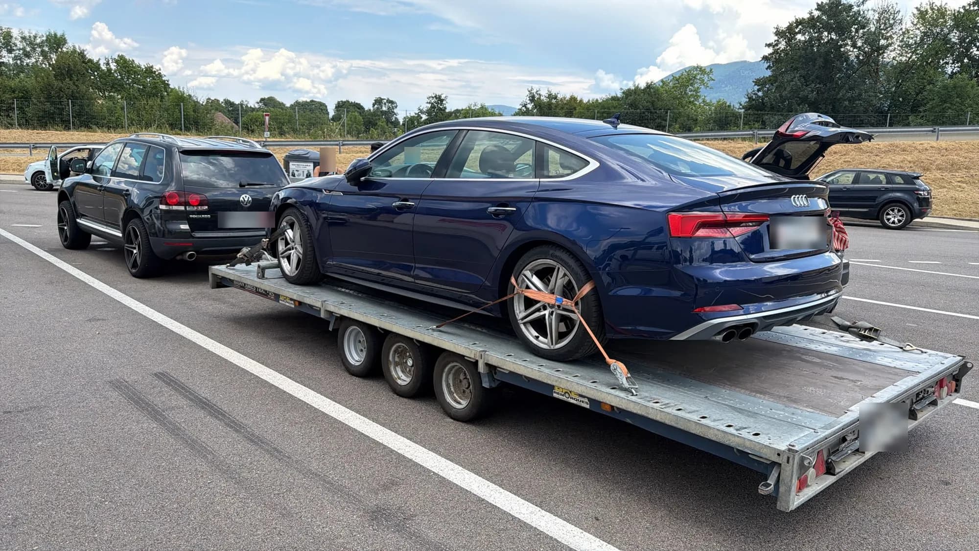 Berline Audi bleu foncé sanglée sur une remorque porte-voiture à trois essieux, tractée par un SUV noir sur un parking ensoleillé