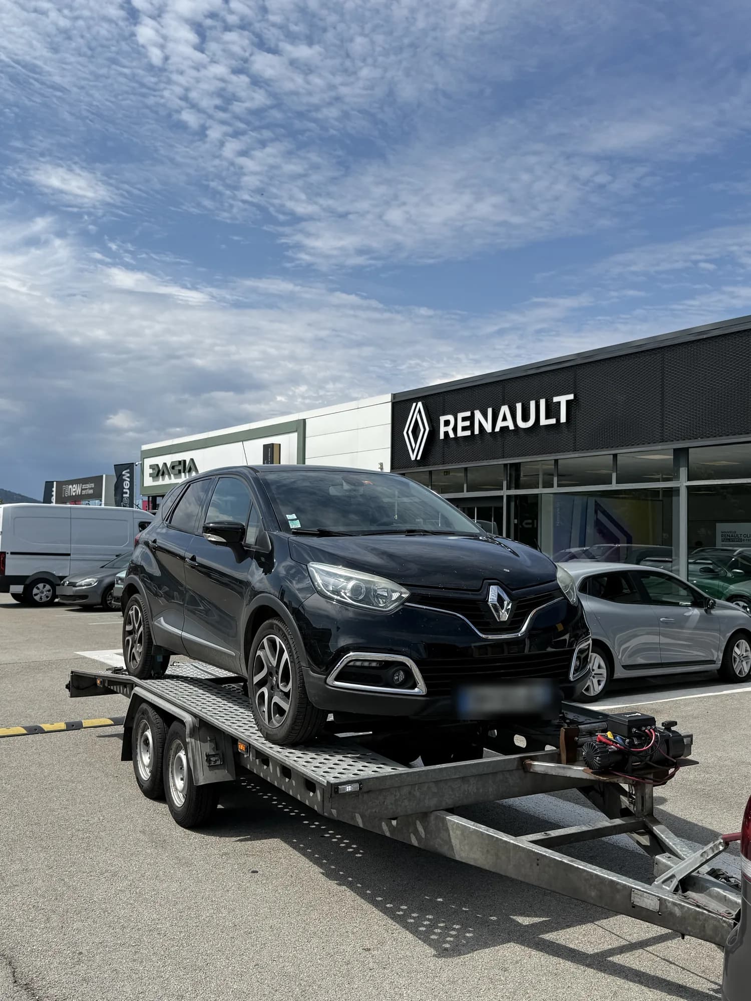 SUV compact Renault noir chargé sur une remorque porte-voiture devant une concession Renault et Dacia, sous un ciel nuageux