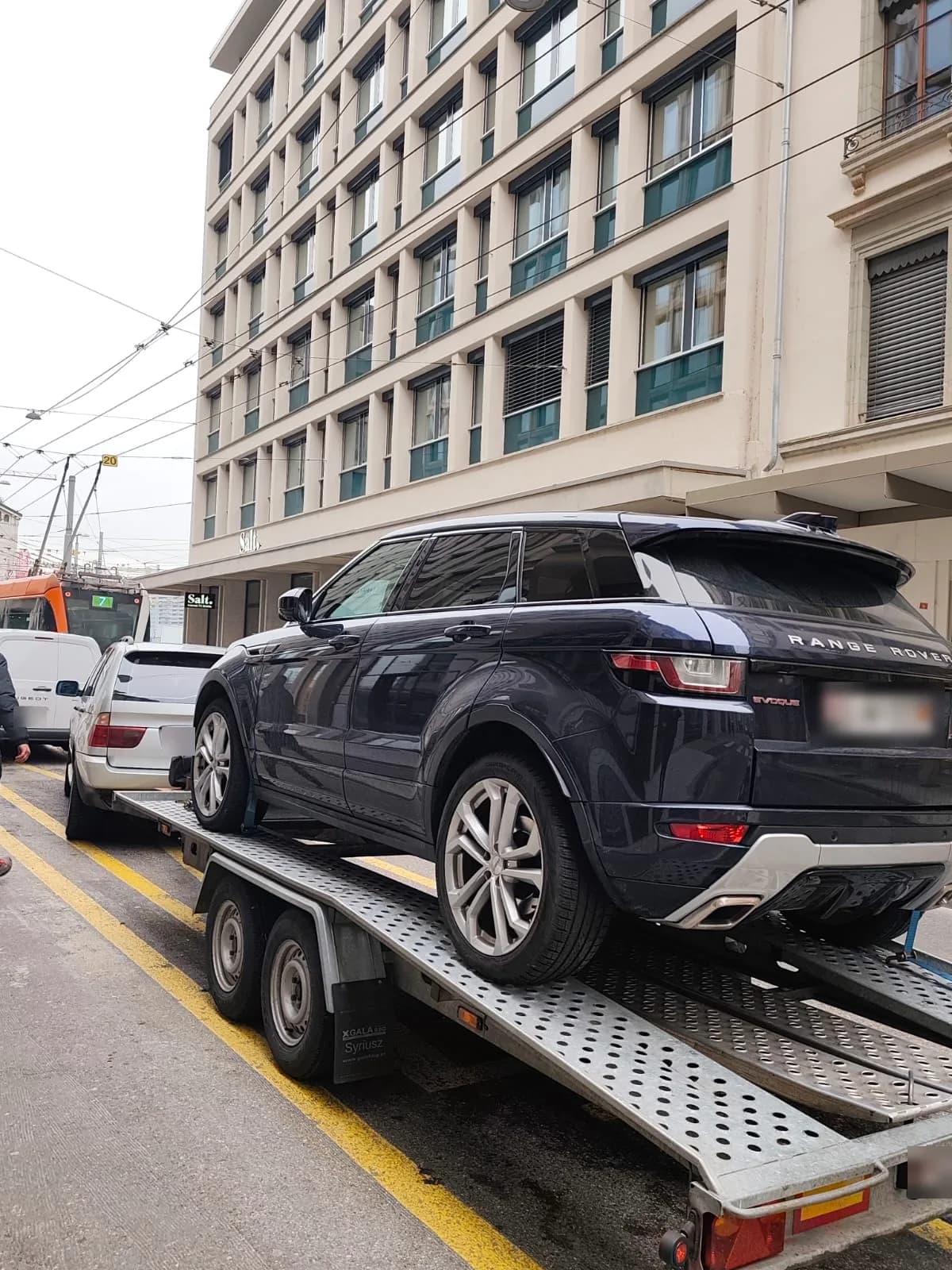 SUV Range Rover bleu nuit sanglé sur une remorque porte-voiture tractée dans une rue de ville, au pied d'un immeuble