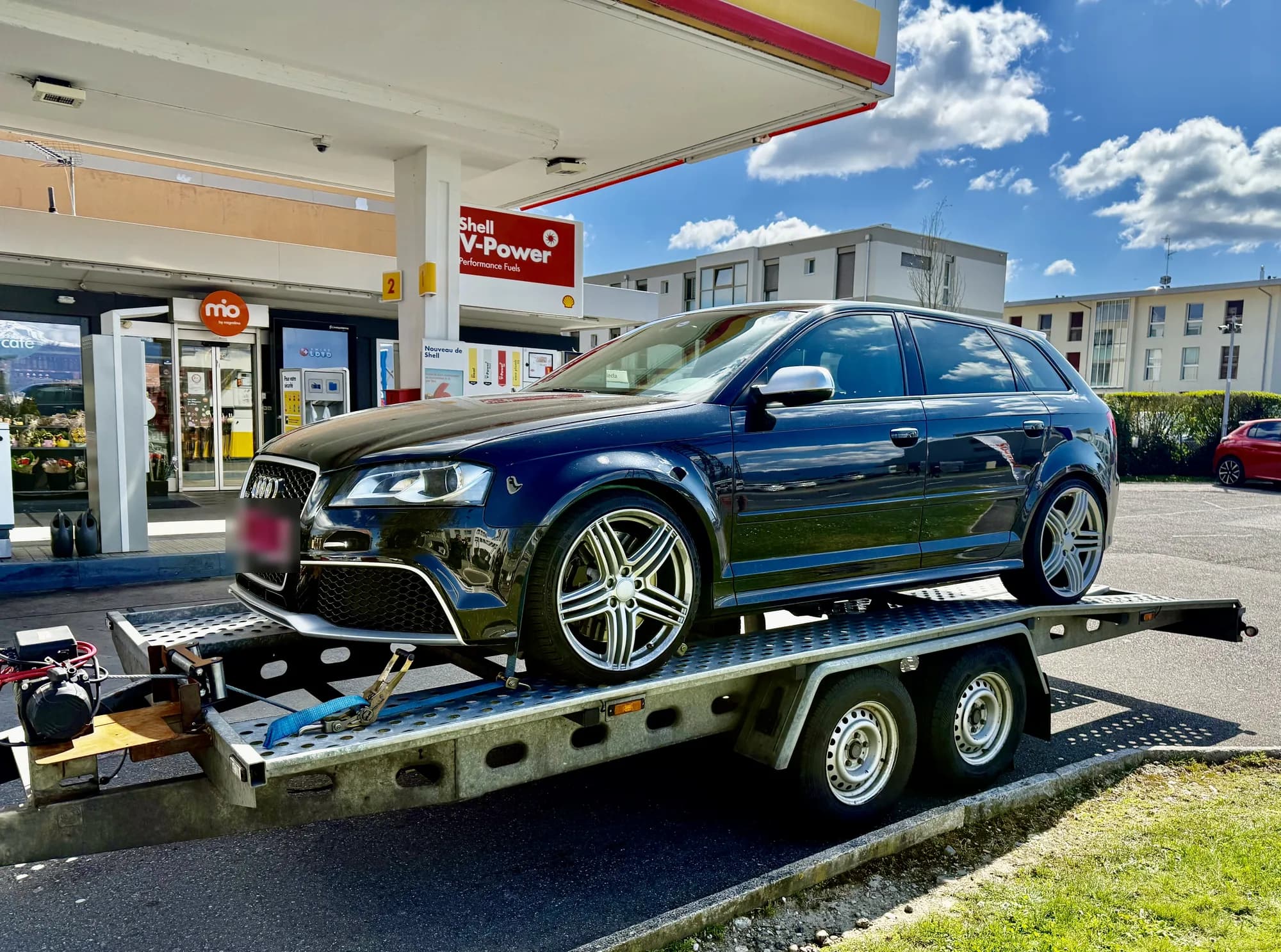 Compacte Audi noire sanglée sur une remorque porte-voiture à plateau perforé, dans une station-service sous un ciel bleu