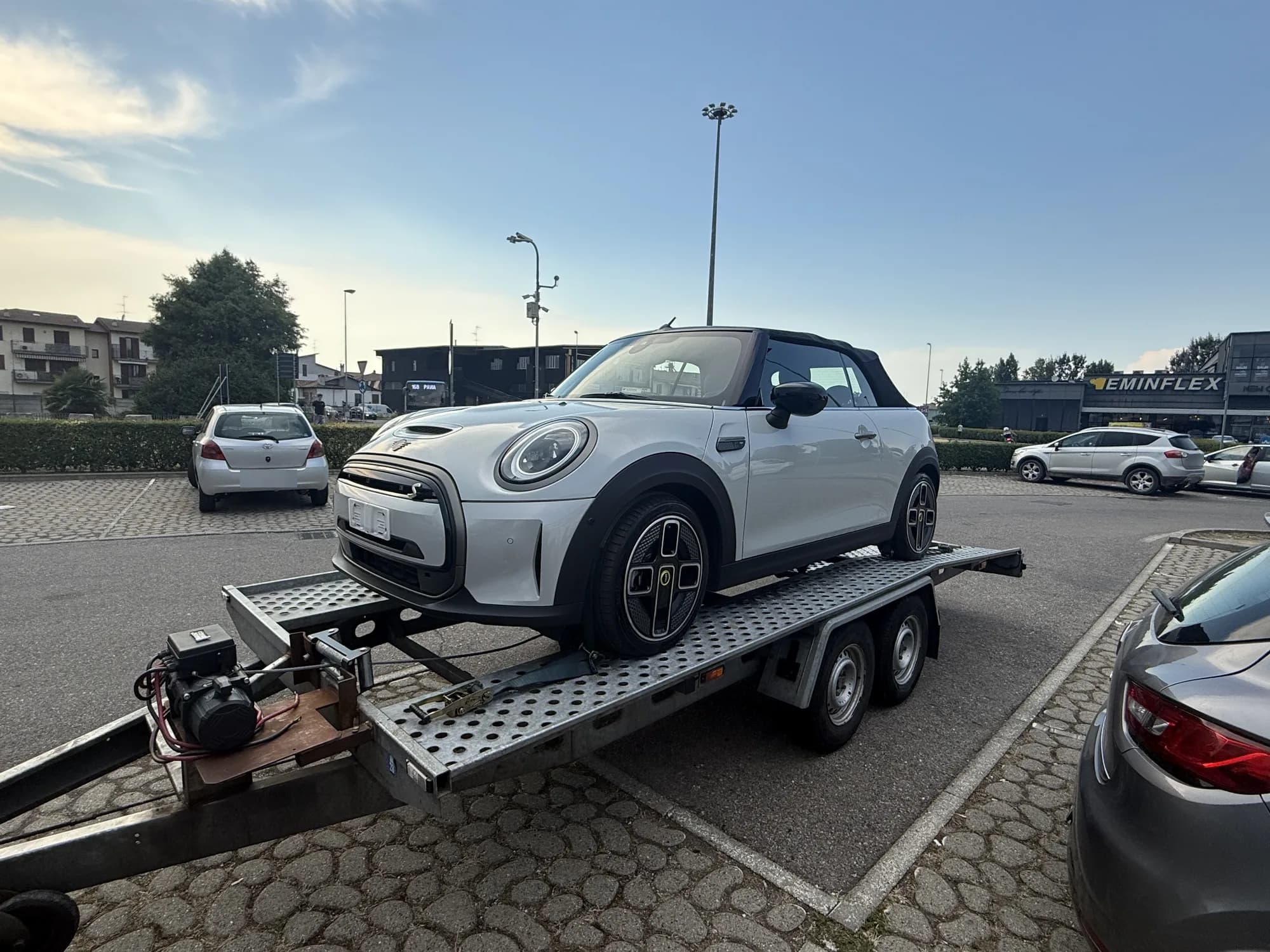 Citadine cabriolet blanche à capote noire chargée sur une remorque porte-voiture à plateau perforé, sur un parking pavé