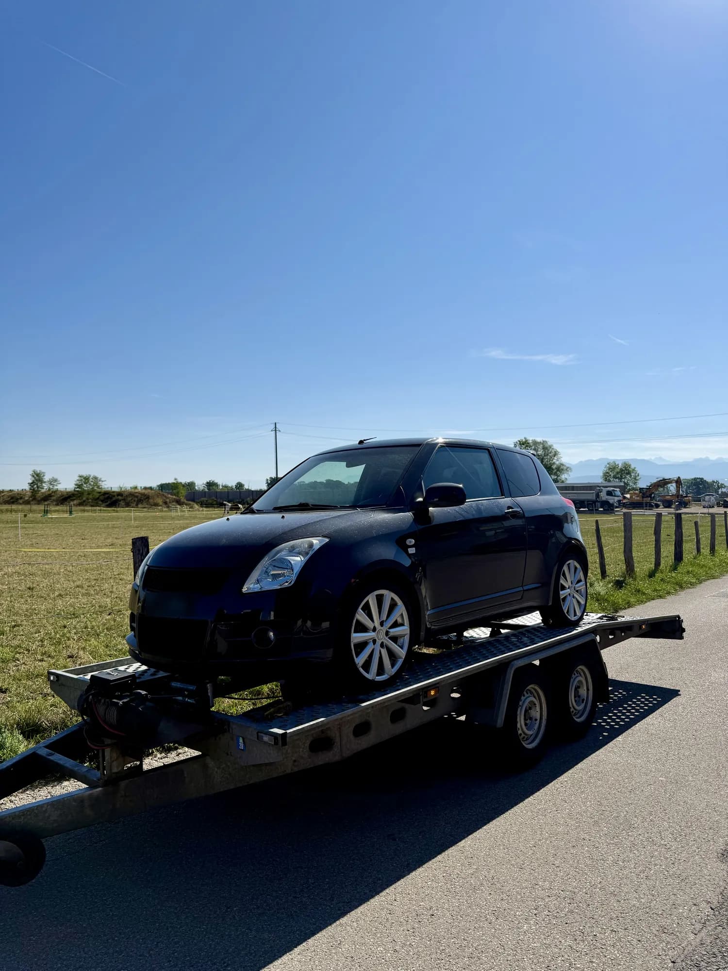 Citadine noire chargée sur une remorque porte-voiture à double essieu, au bord d'une route de campagne sous un ciel bleu