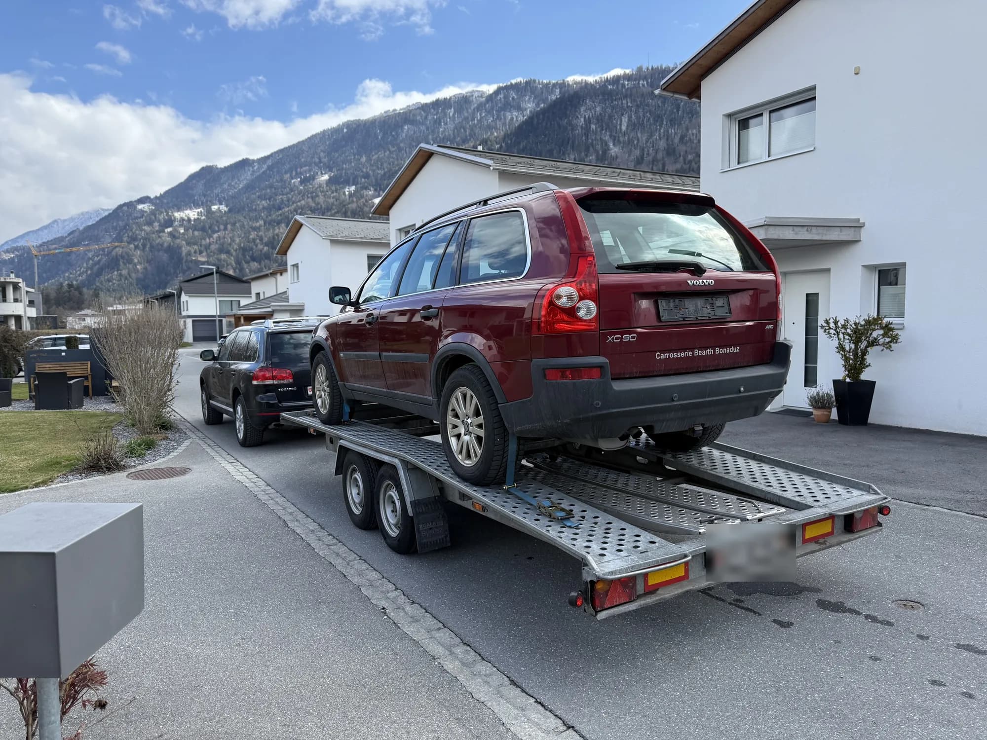 SUV Volvo rouge sanglé sur une remorque porte-voiture tractée par un SUV noir, dans un quartier résidentiel au pied des montagnes