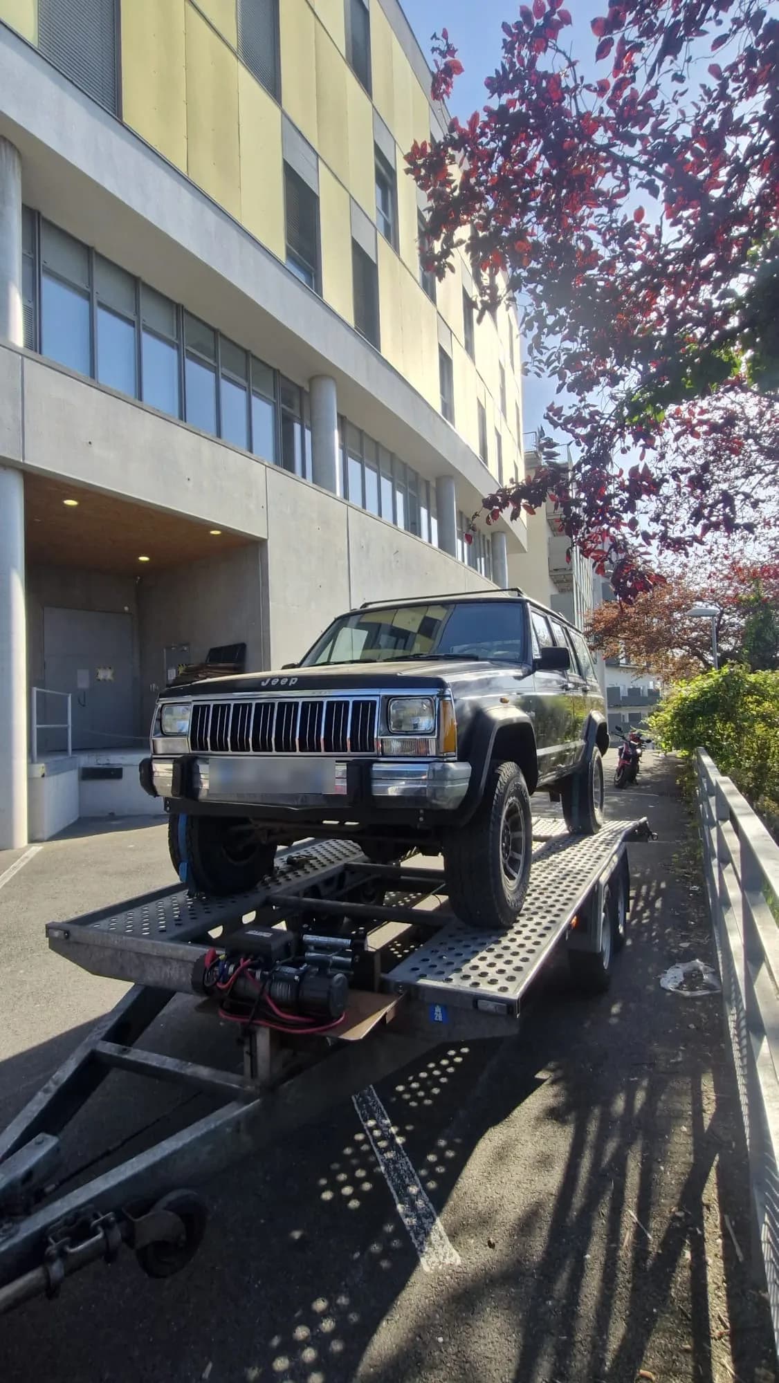 4x4 Jeep noir chargé sur une remorque porte-voiture avec treuil, devant un immeuble moderne en ville, au soleil