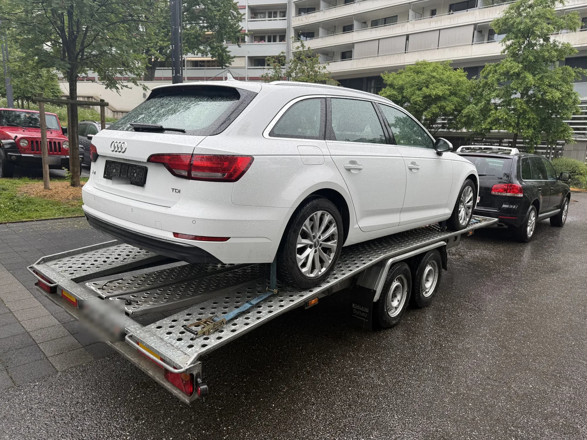 Break Audi blanc sur une remorque porte-voiture perforée tractée par un SUV noir, sur une chaussée mouillée au pied d'immeubles