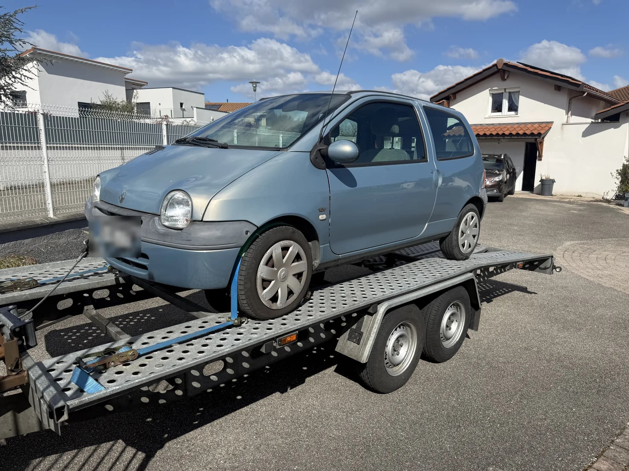 Citadine Renault bleu clair sanglée sur une remorque porte-voiture à double essieu, dans un quartier résidentiel ensoleillé