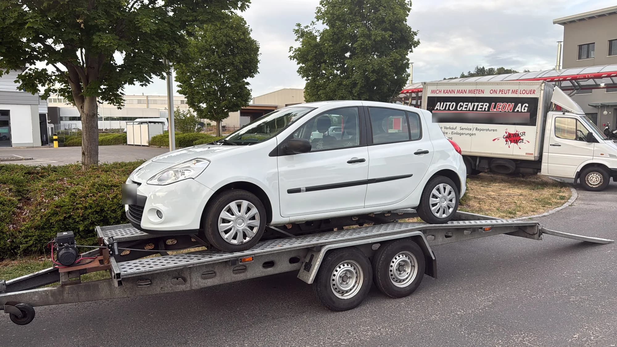 Citadine blanche chargée sur une remorque porte-voiture à double essieu avec treuil, sur le parking d'un garage