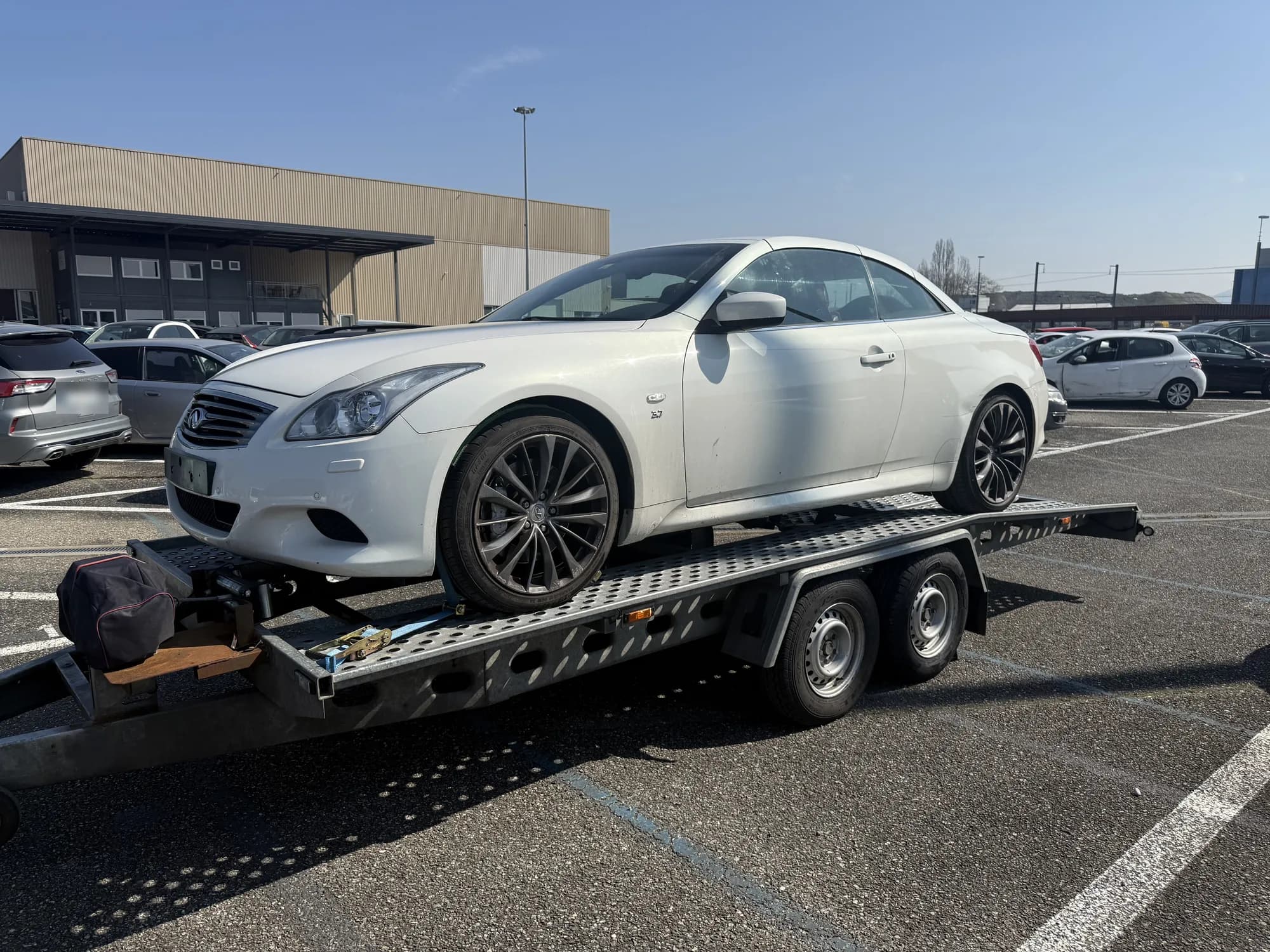 Coupé blanc chargé sur une remorque porte-voiture à double essieu, sur un parking ensoleillé devant un bâtiment industriel