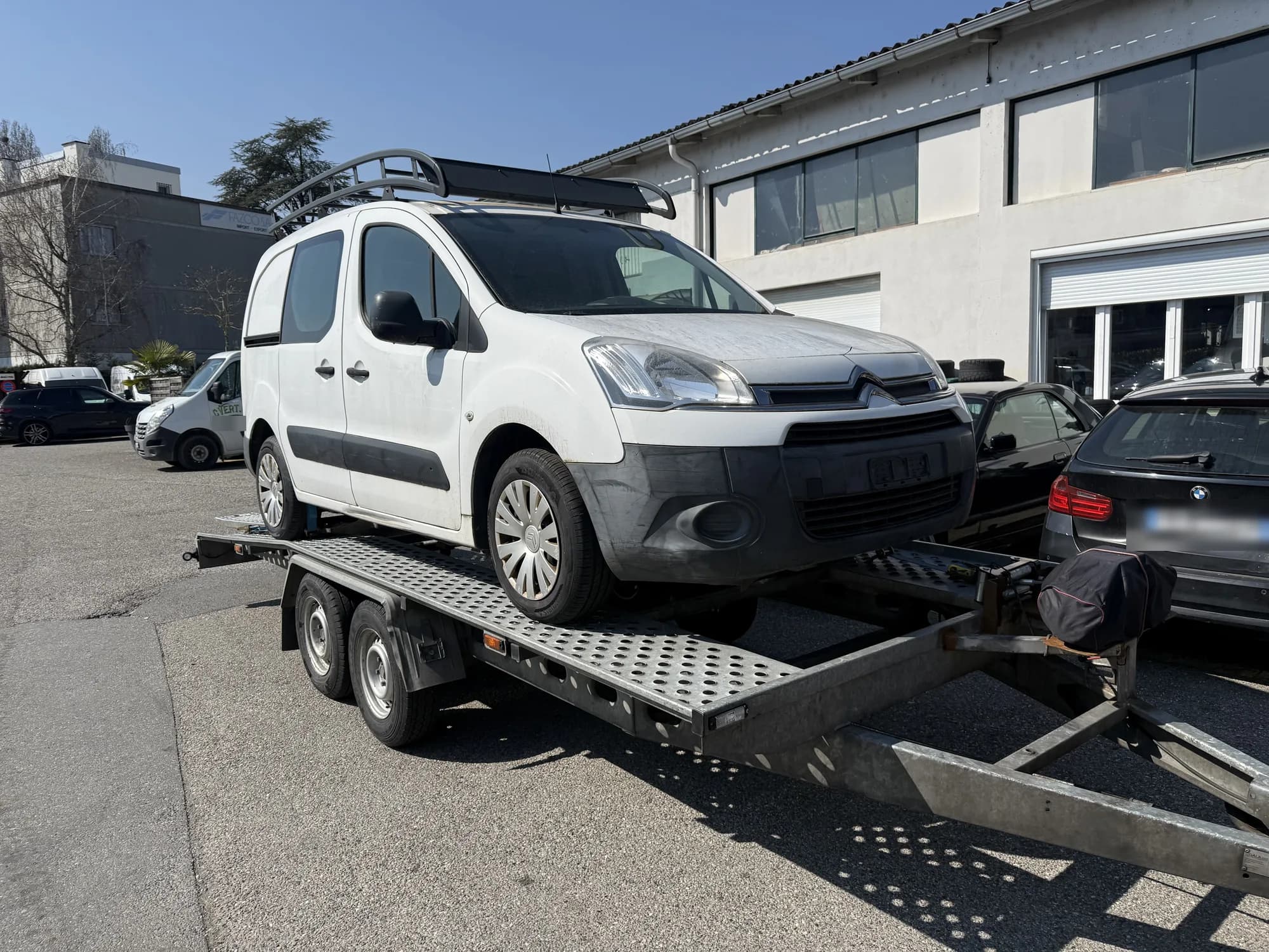 Fourgonnette Citroën blanche à galerie de toit chargée sur une remorque porte-voiture, sur le parking ensoleillé d'un bâtiment industriel