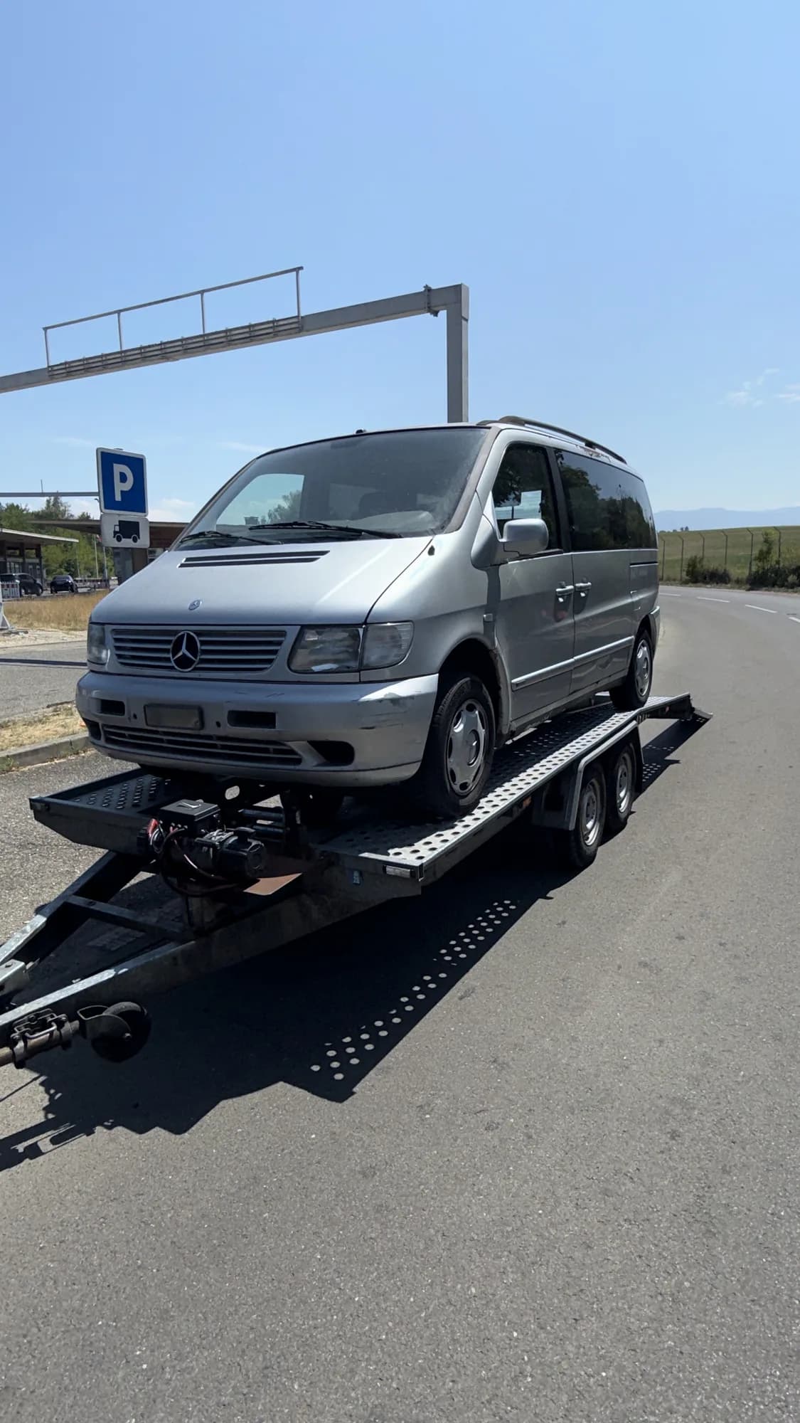 Monospace gris argenté chargé sur une remorque porte-voiture à double essieu, au bord d'une route près d'un parking
