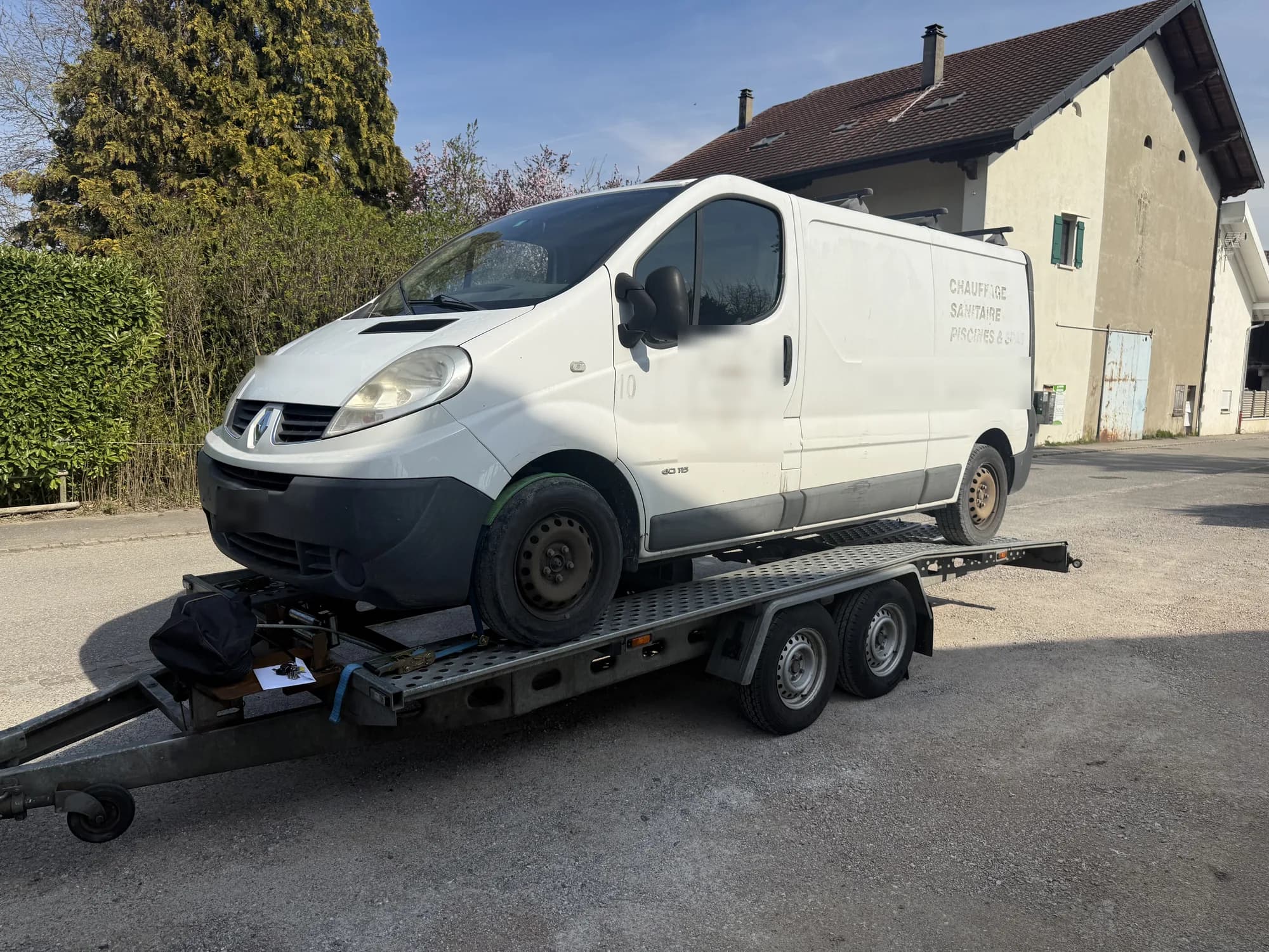 Utilitaire Renault blanc chargé sur une remorque porte-voiture à double essieu, devant une maison de village ensoleillée