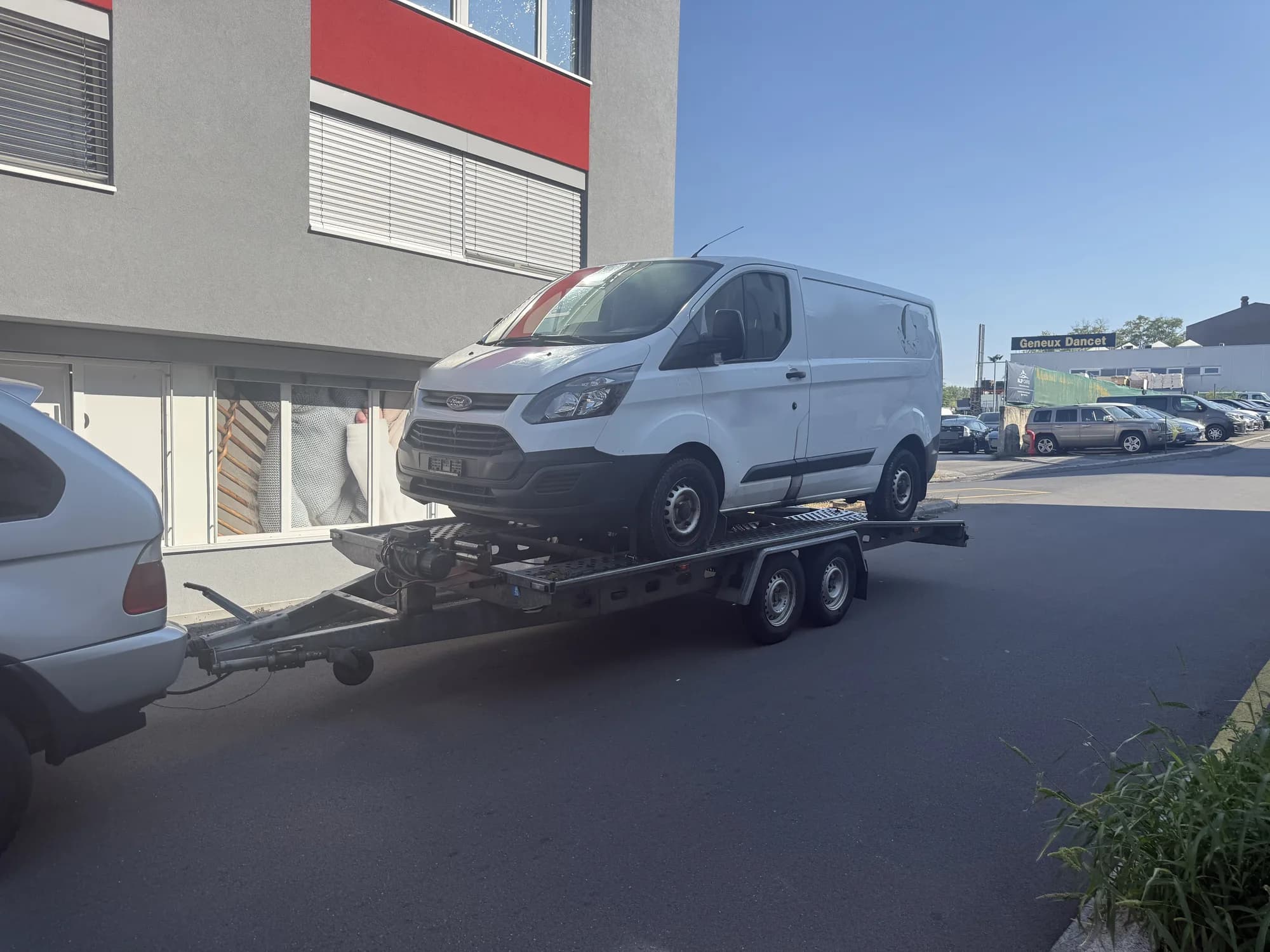 Fourgon utilitaire blanc chargé sur une remorque porte-voiture tractée par un SUV gris, dans une rue devant un immeuble