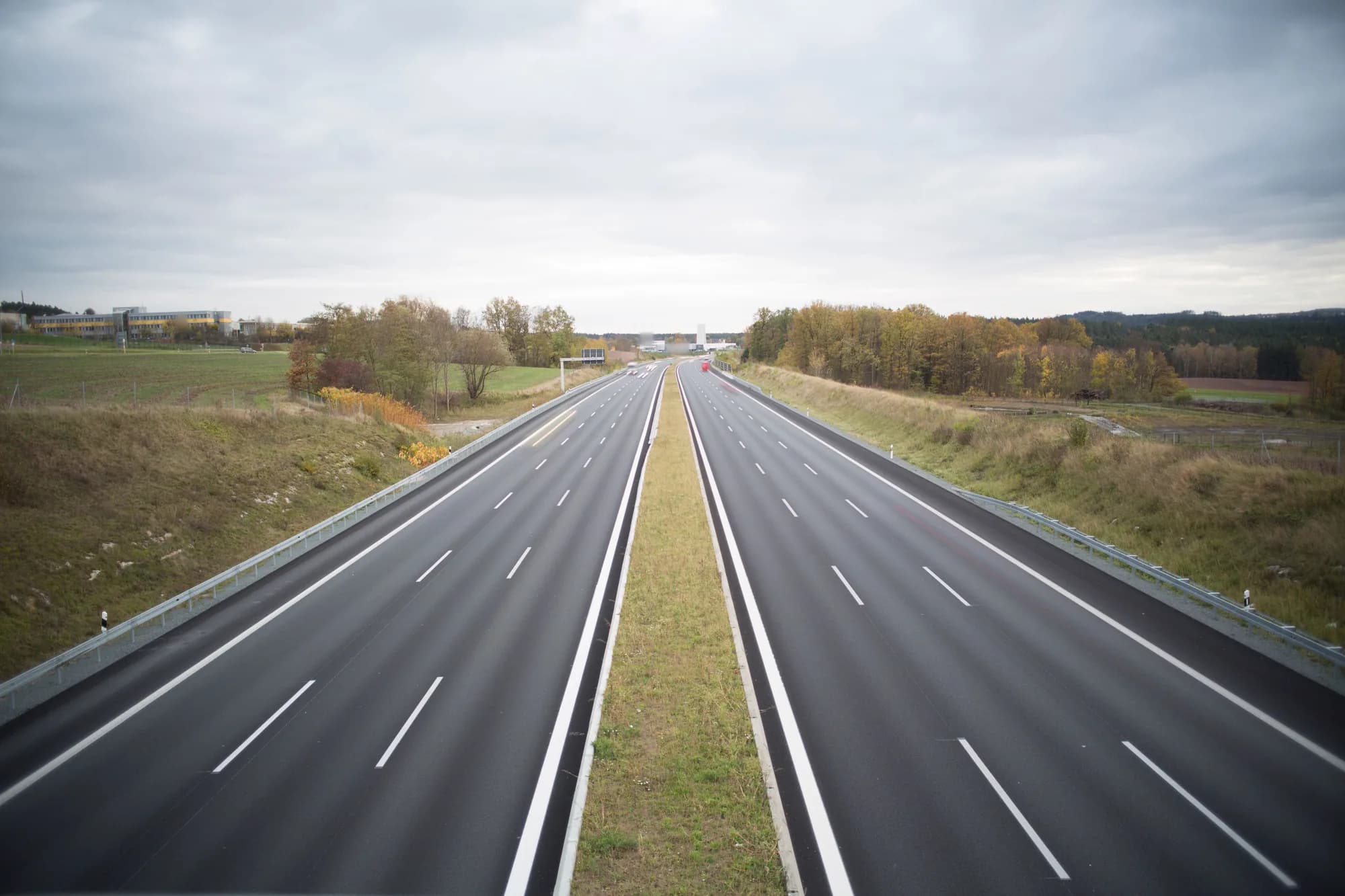 Large autoroute presque vide qui file vers l'horizon, entre champs et bois d'automne, sous un ciel gris