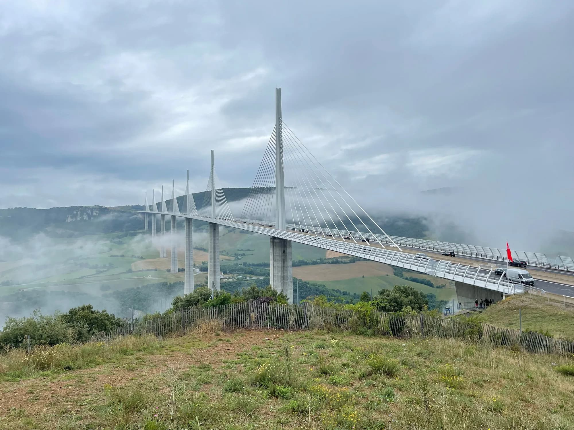 Viaduc de Millau et ses haubans blancs, parcouru par quelques voitures, au-dessus d'une vallée verte traversée de nappes de brume