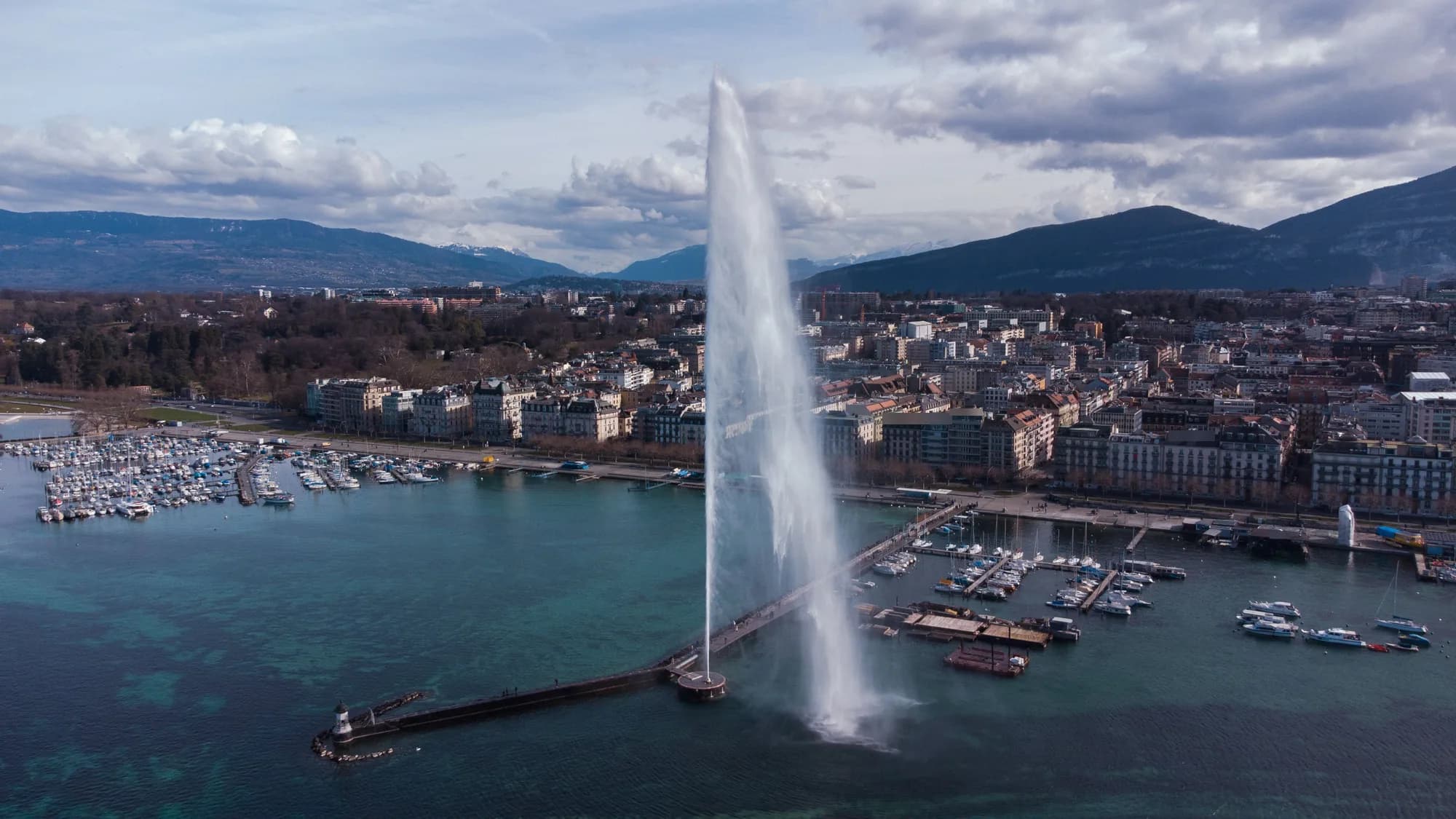 Vue aérienne du Jet d'eau de Genève, de sa jetée, des ports et des quais devant les montagnes, ciel nuageux