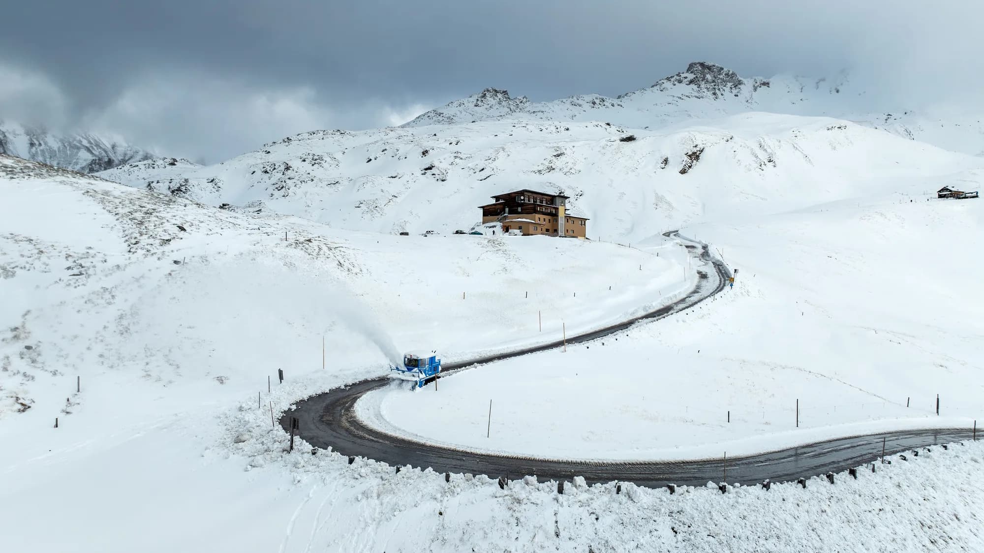 Route de montagne enneigée en lacets où une fraiseuse à neige bleue dégage la chaussée, sous un ciel gris chargé
