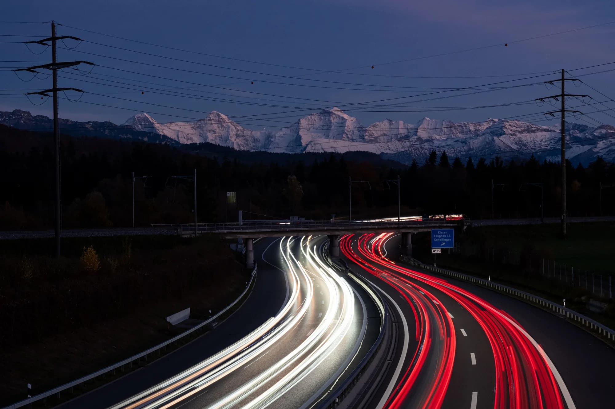 Autoroute à l'heure bleue, traînées lumineuses blanches et rouges sous un pont, sommets enneigés des Alpes bernoises derrière des lignes électriques